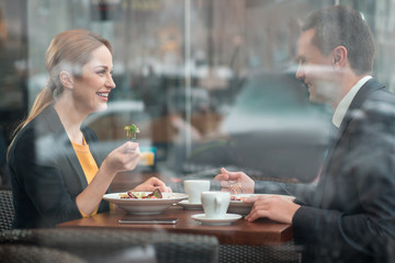 Side view smiling female and outgoing businessman eating portion of salad while sitting at desk in cafe. Lunch concept