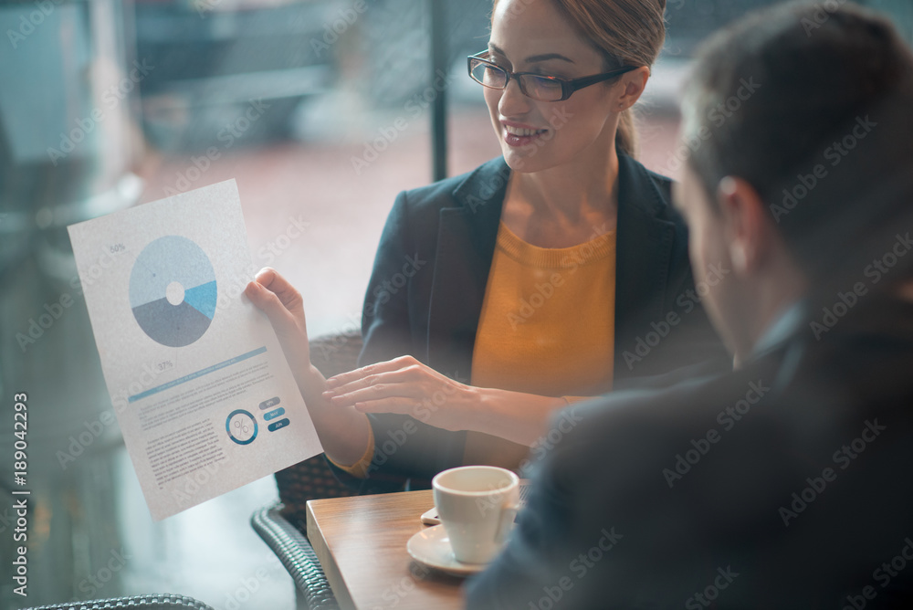 Poster outgoing girl showing scheme to partner. he looking at it. they sitting at table. presentation conce