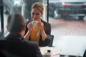 Portrait of cheerful female tasting cup of coffee while talking with colleague. They sitting at table in restaurant. Business break concept. Copy space