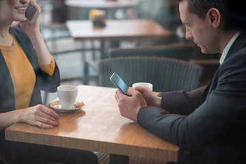 Smiling man watching at monitor of modern phone while situating at desk near colleague. Female speaking by appliance. Rest and work concept