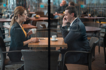 Side view outgoing male talking by phone while situating at desk opposite female colleague. They drinking cup of coffee in restaurant. Break concept