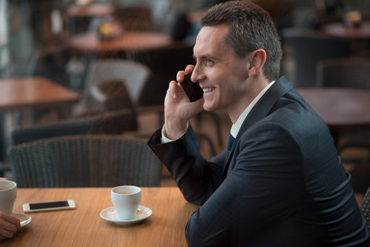 Side view smiling businessman telling by mobile while sitting at table in cafe. Communication and work concept