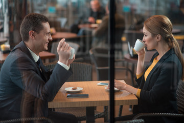 Side view smiling man telling with happy female partner while tasting mug of coffee. They sitting at table. Leisure concept