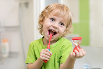 Happy child boy brushing teeth near mirror in bathroom. He is monitoring lasting of cleaning action with hourglass.