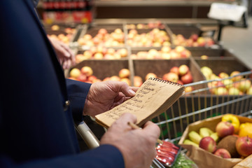 Close up of senior man holding shopping list while grocery shopping in supermarket, fruits and...