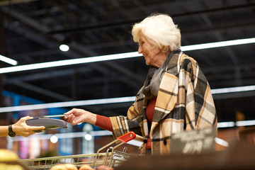 Side view portrait of modern senior woman paying with NFC payment via smartphone  and smiling happily while buying groceries in supermarket, copy space
