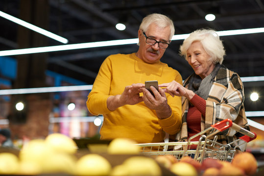 Portrait of cheerful senior couple grocery shopping in supermarket, using smartphone to read shopping list and calculate prices while standing by fruits and vegetables isle, copy space