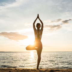 Silhouette of a woman practicing the tree yoga pose on a beach at sunset during summer vacation