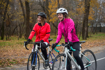 Obraz premium Two Young Smiling Female Cyclists with Road Bicycles Resting and in Park in Cold Autumn Day. Healthy Lifestyle.