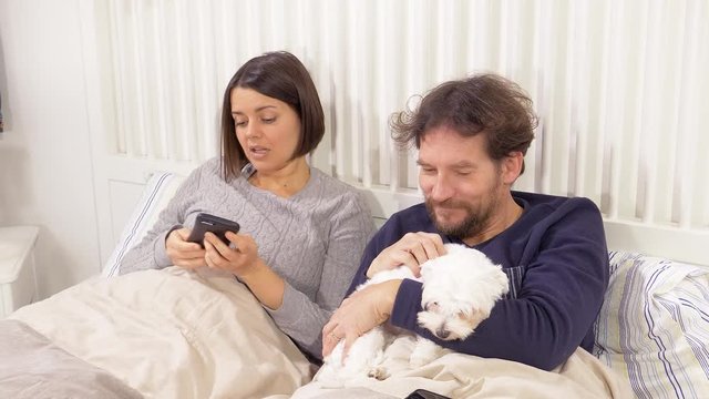 Couple in pajamas in bed taking selfie with puppy dog