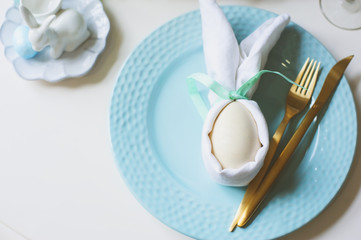 Fototapeta na wymiar easter festive table. Guest dining place decorated with bunny napkin and egg, golden metalic cutlery and blue plate