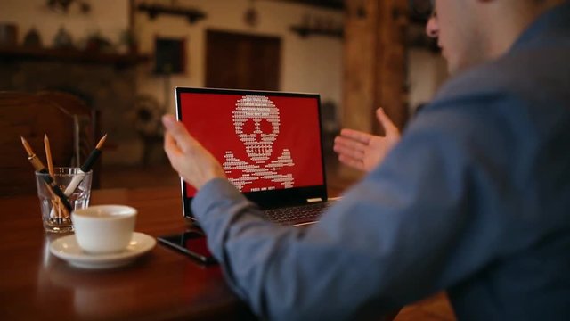 Man turns on a laptop waits for loading computer and finds out it is infected by a ransomware spyware virus that is asking for money to retrieve data files. Scary red skull and crossbones on screen.