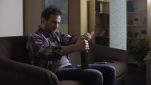 Man sitting on couch in dark room, trying to open beer bottle with fork, despair