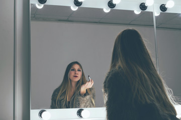 Young blonde dancer in a sexual fur coat checks her makeup before performing at the party. Concept of female narcissism