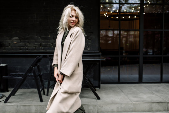 Portrait of beautiful young stylish blonde woman wearing beige coat and walking through the city streets. Trendy casual outfit. Street fashion.
