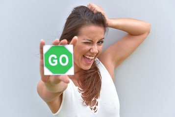 A young girl stands in front of a grey cement wall. She holds up a green card to the camera with the word go on it.