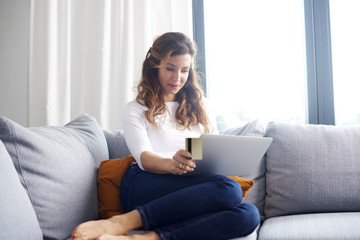 Banking from home is comfy. A woman sitting on sofa and looking thoughtful while shopping online with her credit card.