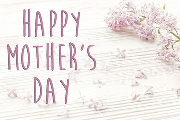 happy mother's day text sign, simple greeting card. hello spring image. beautiful tender lilac flowers and petals in light on white wooden rustic background top view. happy mothers day