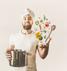 Young cook man with hat and beard holding pan and spoon with flying vegetables on white background....