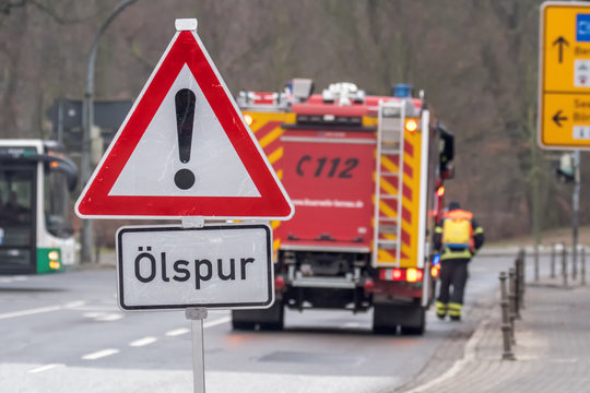 A signal sign indicates a spilled oil on the road. The fire department secures the danger spot. The sign says in German: oil track. Concept: road problems or safety