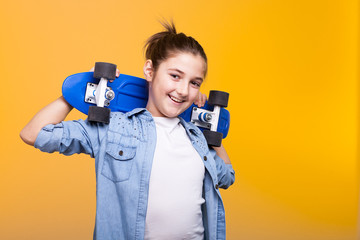 Happy cool teenager girl with a blue skateboard in hands on yellow background