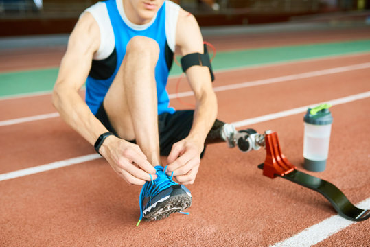 Unrecognizable Amputee Athlete Tying Shoe Laces Sitting On Running Track In Modern Indoor Stadium, Copy Space