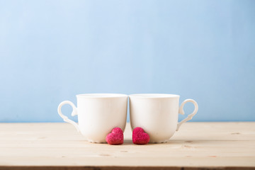 Obraz na płótnie Canvas Valentines day concept, mugs cups of coffee or tea for two lovers honeymoon wedding morning surprise breakfast, pink heart sugar candies, blue background pastel colors copy space, light wooden table 
