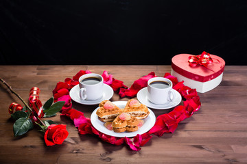 On the wooden table, inside the heart of the rose petals are two cups of coffee and a plate with biscuits. Outside, a rose flower and a box with a gift. The middle plan