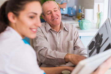 Obraz premium The old happy man sitting at the table with the young cheerful doctor and they reviewing photo album in the kitchen