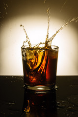 Glass of whiskey with spray from the thrown ice / In the picture there is a glass of whiskey with ice, standing on a table filled with liquid
