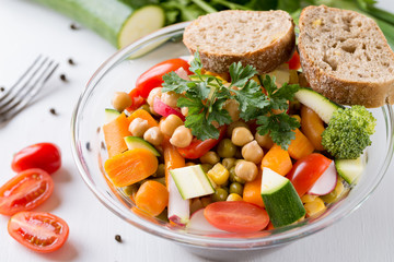 Vegetable salad in glass bowl with broccoli and tomatoes on white wooden table.