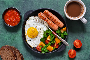 Good morning! Breakfast with sausage, egg, salad and tomato sauce with a cup of coffee on a green concrete background.