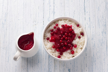 Rice pudding with cranberry jam and fresh cranberries in a white bowl on a light blue background, selective focus. Delicious traditional breakfast