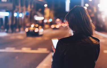 Girl pointing finger on screen smartphone on background illumination bokeh light in night  city, hipster girl using in hands mobile phone, lights auto taxi in glitter street, lifestyle concept
