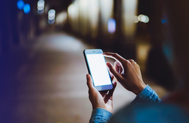 Woman pointing finger on blank screen smartphone on background bokeh light in night atmospheric city, hipster using in hands clean gadget mobile phone, mockup street, online wifi internet concept
