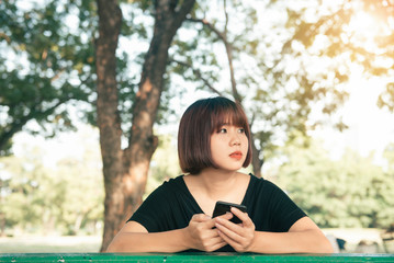 Cute asian woman reading pleasant text message on mobile phone while sitting in park spring day. Asian woman using on smart phone with feeling relax and smiley face. Lifestyle and technology concepts.