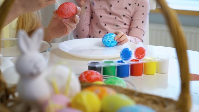 Happy Easter! Young mother and her cute little daughter wearing funny rabbit ears are coloring easter eggs sitting at a festive table with basket and Bunny. Slow Motion