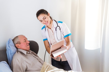 Obraz premium The old, ill man lying on a bed and the young cheerful doctor standing near him and showing the tablet him in the room