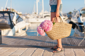 Traveler is standing in yachts port near sea with straw bag and pink flowers hydrangea. Girl has...