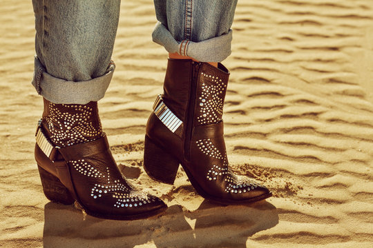 Close up photo of black leather cowboy ankle boots with decorative metallic elements. Model walking on the sand. Copy, empty space for text