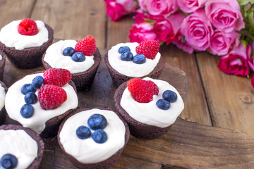 Chocolate muffins with raspberries and blueberries, and white cheese cream, on a brown wooden board. A bouquet of fresh pink roses for lovers. Homemade pastries for breakfast. Free space for text or p