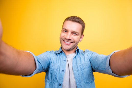 Self portrait of attractive, cute, smiling man with bristle, stubble in jeans shirt, shooting selfie with two hands on front camera over yellow background