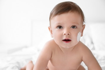 Cute baby with body cream on cheek against light background