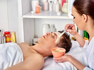 Obraz na płótnie Canvas Mud facial mask of man in spa salon. Massage with mud from full face. Male lying spa bed. Beautician with bowl therapeutic procedure . Anti-aging cosmetic dead sea mud mask.