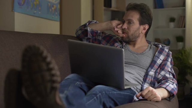 Guy sitting on sofa, looking at laptop, angry with received news, bitterness