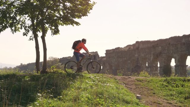 Cyclist with bike reads map for direction in front of ancient roman aqueduct in a park. Young attractive athletic man with orange sportswear and backpack in parco degli acquedotti in Rome under a tree