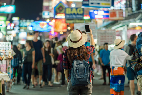 Back side of Young Asian traveling women taking photo in Khaosan Road walking street at night in Bangkok, Thailand, traveler and tourist concept