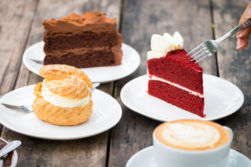 Close-up of woman's hand holding fork to eating Red velvet cake, chocolate cake, Shu cream (Japanese cream puff) and hot coffee on wooden table in outdoor garden. Concept sweets enjoy eating.
