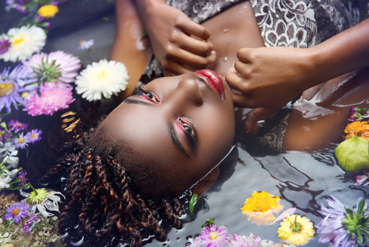 African woman with bright make up lies in the water with flowers close-up upside-down