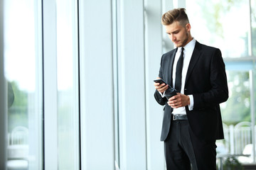 Young businessman standing in an office near the window and talking on a cell phone.